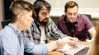 three male students talk to each other while looking at laptop screen